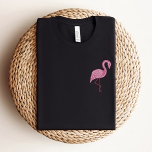 May include: Black t-shirt with a pink flamingo graphic. The t-shirt is folded and displayed on a woven straw mat.