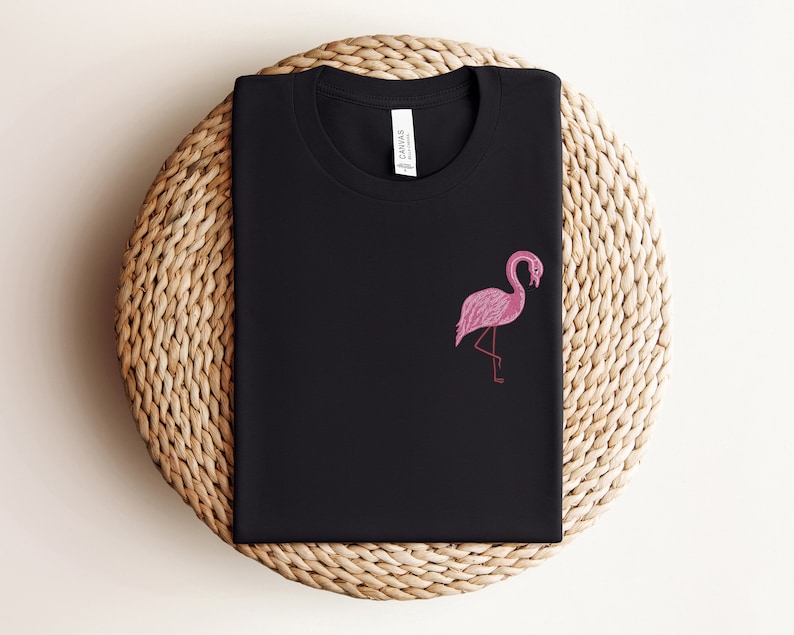 May include: Black t-shirt with a pink flamingo graphic. The t-shirt is folded and displayed on a woven straw mat.