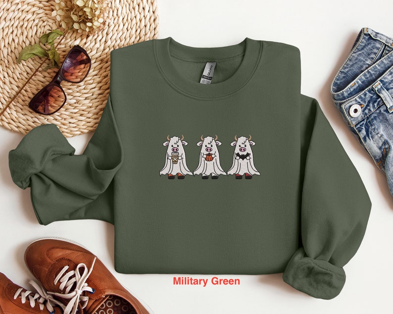 K&ouml;nnte beinhalten: Ein olivgr&uuml;nes Sweatshirt mit Rundhalsausschnitt und langen &Auml;rmeln. Das Sweatshirt zeigt ein Motiv mit drei wei&szlig;en K&uuml;hen, die als Geister verkleidet sind und einen K&uuml;rbis, eine Kaffeetasse und einen Mais-S&uuml;&szlig;igkeiten halten. Der Text "Milit&auml;rgr&uuml;n" ist unter dem Motiv gedruckt.