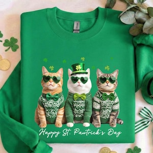 May include: Green sweatshirt featuring three cats wearing green heart-shaped sunglasses, bow ties, and sweaters. The cats are adorned with St. Patrick's Day themed accessories. The text "Happy St. Pawtrick's Day" is printed at the bottom.