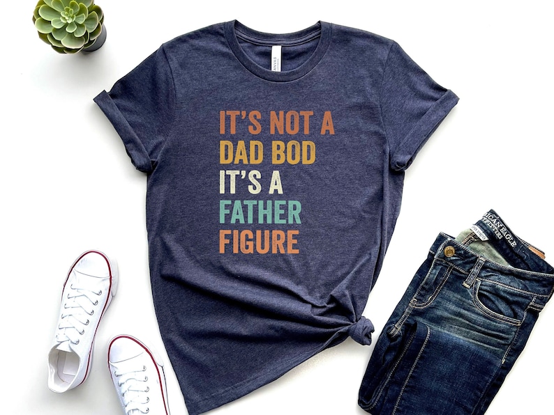 Pu&ograve; includere: Una maglietta blu scuro con il testo "It's not a dad bod it's a father figure" in un font retr&ograve; con accenti colorati.