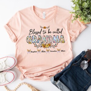 May include: A peach-colored t-shirt with the text "Blessed to be called GRANDMA" in floral letters. The text "Sophia, Alex, Brandon, Olive" is printed below the main text.