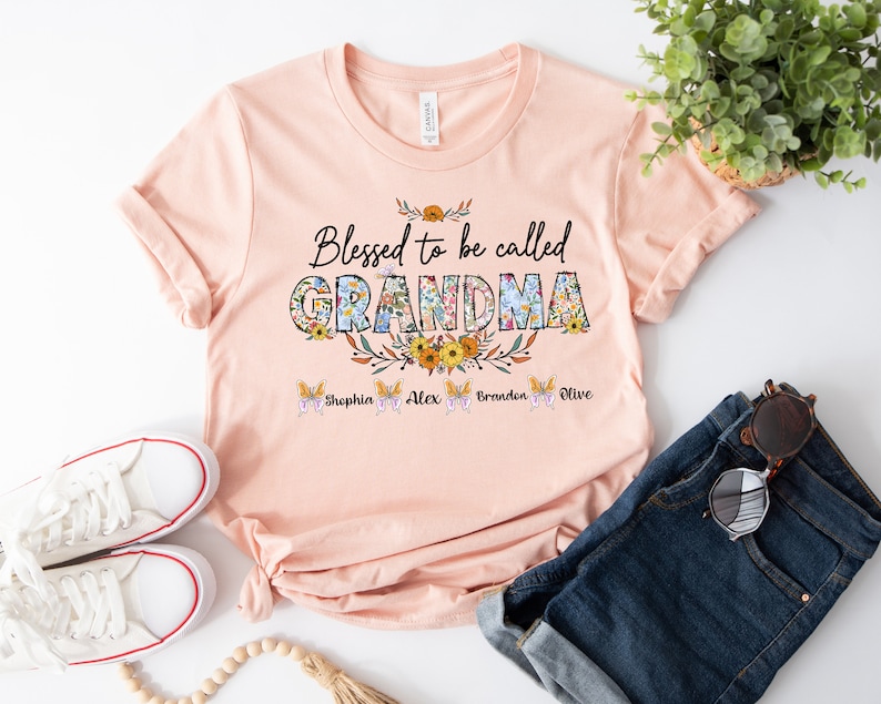 May include: A peach-colored t-shirt with the text "Blessed to be called GRANDMA" in floral letters. The text "Sophia, Alex, Brandon, Olive" is printed below the main text.