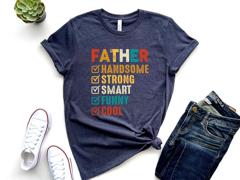 K&ouml;nnte beinhalten: Ein dunkelblaues T-Shirt mit dem Text "FATHER" in Wei&szlig; und einer Checkliste mit positiven Eigenschaften in bunter Schrift. Die Eigenschaften sind "handsome", "strong", "smart", "funny" und "cool".