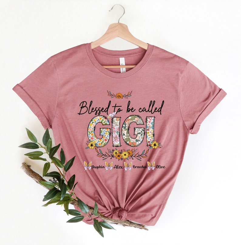 May include: A pink t-shirt with the text "Blessed to be called Gigi" in black lettering. The word Gigi is decorated with floral patterns. Below the text are the names "Sophia, Alex, Branden, Olive" in black lettering.