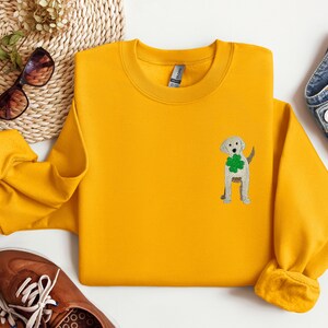 May include: A yellow sweatshirt with a white embroidered dog wearing a green shamrock. The dog is standing on its hind legs and has its tail curled around its back.