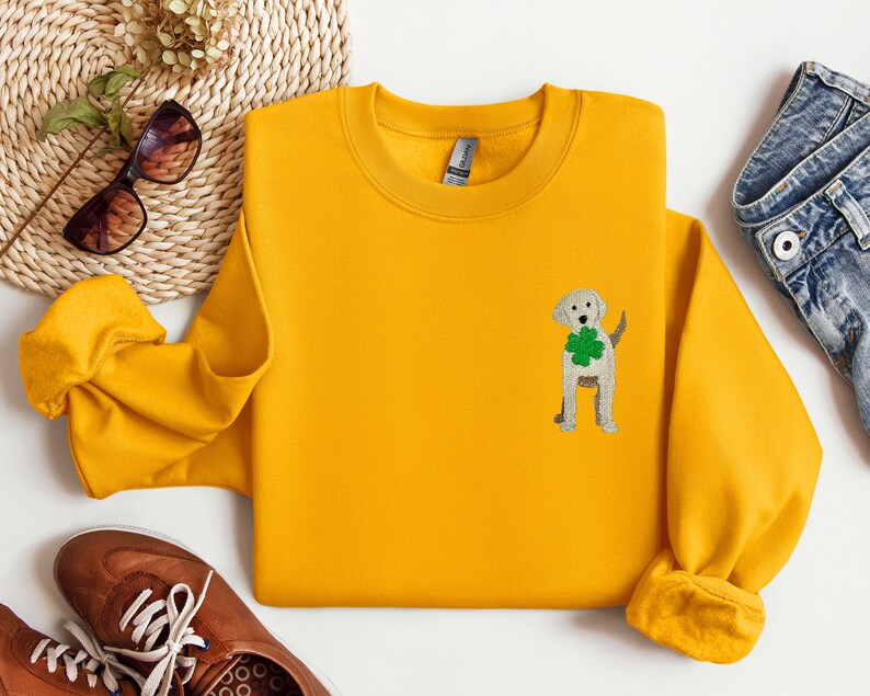 May include: A yellow sweatshirt with a white embroidered dog wearing a green shamrock. The dog is standing on its hind legs and has its tail curled around its back.