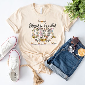 May include: A beige t-shirt with the text "Blessed to be called GIGI" in black lettering. The letters are decorated with floral patterns. Below the text are four butterflies with the names "Sophia", "Alex", "Brandon", and "Olive" written on them.