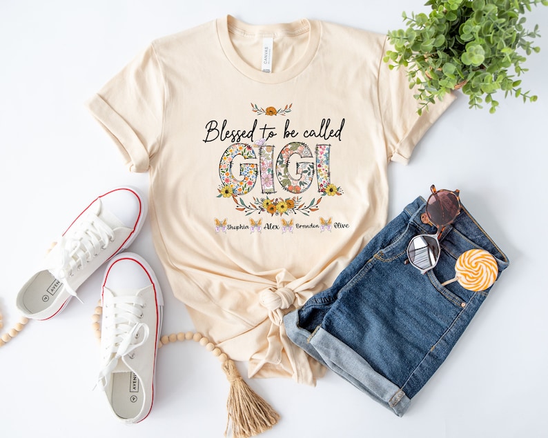 May include: A beige t-shirt with the text "Blessed to be called GIGI" in black lettering. The letters are decorated with floral patterns. Below the text are four butterflies with the names "Sophia", "Alex", "Brandon", and "Olive" written on them.