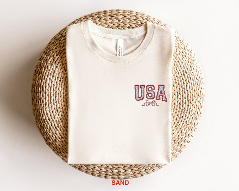 Puede incluir: Una camiseta de color arena con un gr&aacute;fico "USA" rojo, blanco y azul en la parte delantera. El gr&aacute;fico tiene un estilo vintage, de estilo universitario.