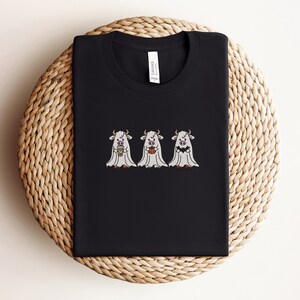 K&ouml;nnte beinhalten: Schwarzes T-Shirt mit drei wei&szlig;en Geisterk&uuml;hen, die eine Tasse Kaffee, einen K&uuml;rbis und einen Mais s&uuml;&szlig; halten. Der Text "Canvas" ist auf der R&uuml;ckseite des Shirts gedruckt.