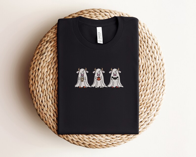 K&ouml;nnte beinhalten: Schwarzes T-Shirt mit drei wei&szlig;en Geisterk&uuml;hen, die eine Tasse Kaffee, einen K&uuml;rbis und einen Mais s&uuml;&szlig; halten. Der Text "Canvas" ist auf der R&uuml;ckseite des Shirts gedruckt.