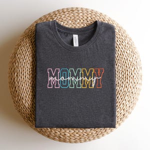 Pode incluir: Uma camiseta cinza escura com a palavra "MOMMY" em letras arco-&iacute;ris. A palavra "mommy" est&aacute; escrita em it&aacute;lico branco dentro das letras.