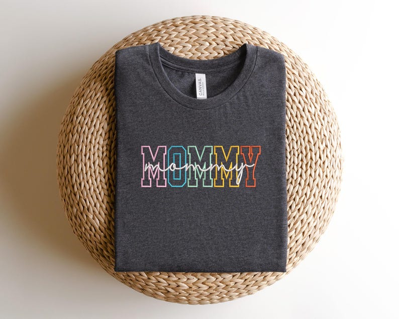 Pode incluir: Uma camiseta cinza escura com a palavra "MOMMY" em letras arco-&iacute;ris. A palavra "mommy" est&aacute; escrita em it&aacute;lico branco dentro das letras.