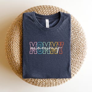 Pode incluir: Uma camiseta cinza escura com a palavra "MOMMY" em letras mai&uacute;sculas arco-&iacute;ris com um "mommy" menor escrito em cursiva abaixo.