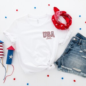 Puede incluir: Una camiseta blanca con la palabra "USA" en letras rojas y azules, un pa&ntilde;uelo rojo, pantalones cortos de mezclilla azul y fuegos artificiales de papel rojo y azul sobre un fondo blanco.