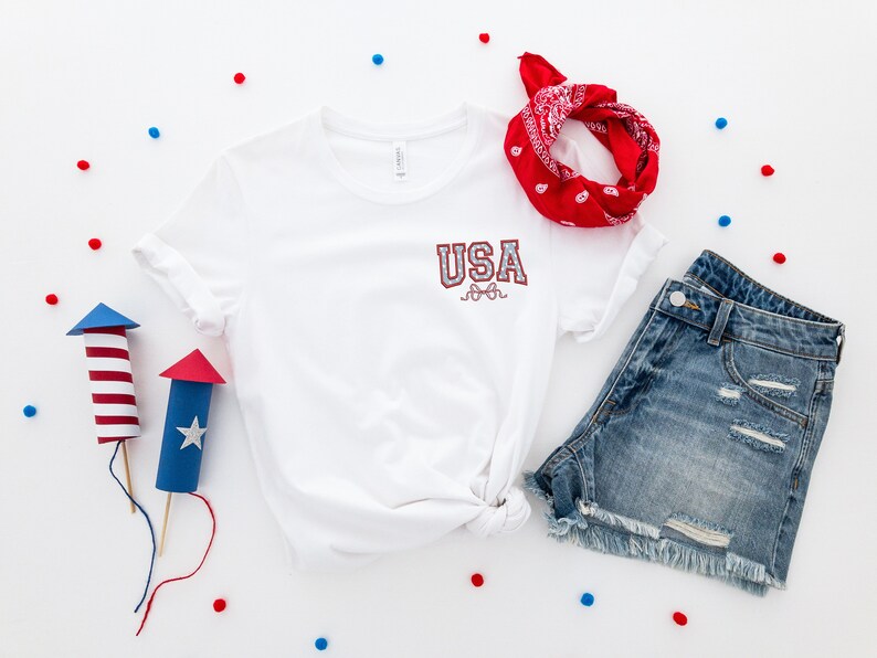 Puede incluir: Una camiseta blanca con la palabra "USA" en letras rojas y azules, un pa&ntilde;uelo rojo, pantalones cortos de mezclilla azul y fuegos artificiales de papel rojo y azul sobre un fondo blanco.