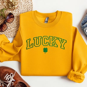 以下が含まれることがあります： 黄色のスウェットシャツに、緑色の縁取りのある文字で「LUCKY」と書いてあり、その下に緑色のクローバーがあります。