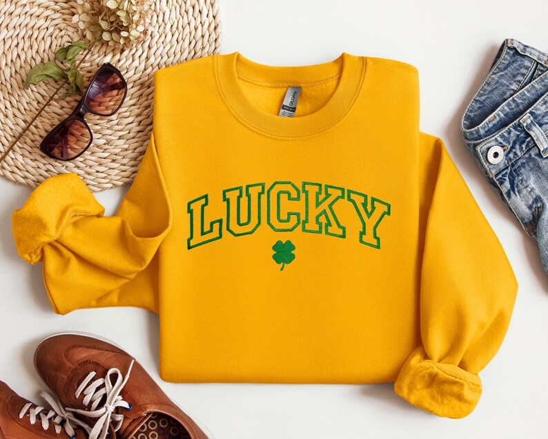 以下が含まれることがあります： 黄色のスウェットシャツに、緑色の縁取りのある文字で「LUCKY」と書いてあり、その下に緑色のクローバーがあります。