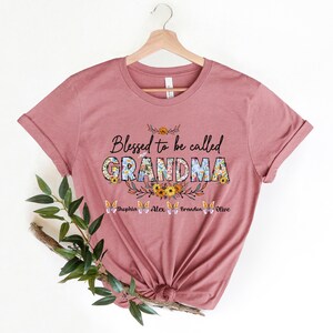 May include: A pink t-shirt with the text "Blessed to be called GRANDMA" in floral letters. The text "Sophia Alex Brandon Olive" is printed below the main text.