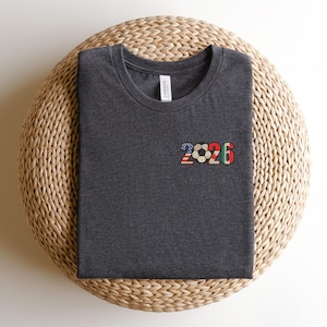 May include: Dark gray t-shirt with a crew neck, featuring the number "2026" in a colorful design. The "2" is in a pattern of the American flag, the soccer ball is in the center, and the "6" is in a green, red, and white pattern.