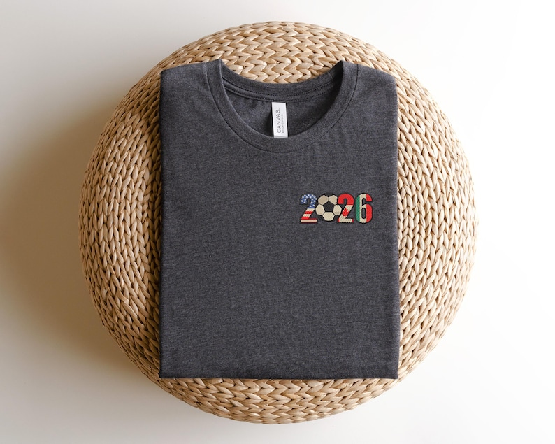 May include: Dark gray t-shirt with a crew neck, featuring the number "2026" in a colorful design. The "2" is in a pattern of the American flag, the soccer ball is in the center, and the "6" is in a green, red, and white pattern.