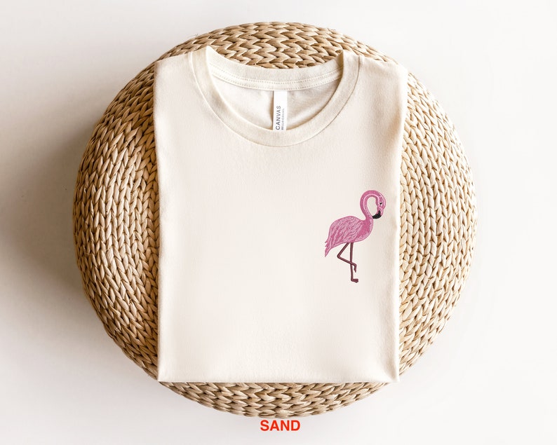 May include: A white t-shirt with a pink flamingo graphic on the front. The shirt is on a woven straw surface. The word "SAND" is printed in red below the shirt.