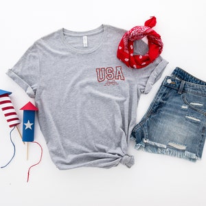 Puede incluir: Una camiseta gris con la palabra "USA" impresa en la parte delantera, una bandana roja, pantalones cortos de mezclilla y dos petardos de papel rojo, blanco y azul.