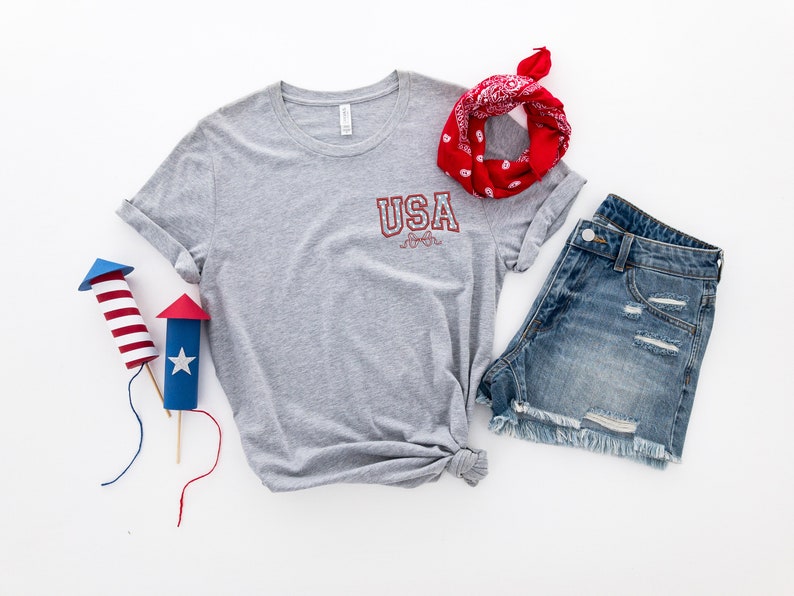 Puede incluir: Una camiseta gris con la palabra "USA" impresa en la parte delantera, una bandana roja, pantalones cortos de mezclilla y dos petardos de papel rojo, blanco y azul.