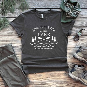 May include: A dark gray t-shirt with white text that reads "Life is better at the LAKE" with a graphic of two crossed paddles and a wavy line representing water. The shirt is laid out on a wooden surface with a pair of green cargo pants, a green bandana, and two brown hiking boots.