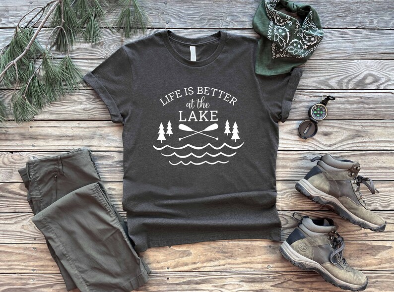 May include: A dark gray t-shirt with white text that reads "Life is better at the LAKE" with a graphic of two crossed paddles and a wavy line representing water. The shirt is laid out on a wooden surface with a pair of green cargo pants, a green bandana, and two brown hiking boots.