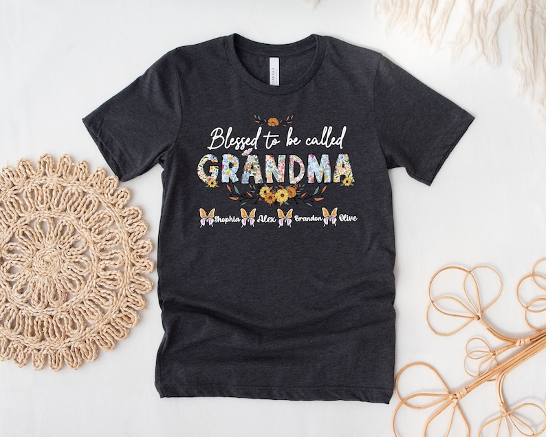 May include: A dark gray t-shirt with the text "Blessed to be called GRANDMA" in white with floral accents. The names "Sophia, Alex, Braeden, Olive" are printed in white below the text.