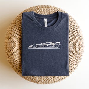 May include: A dark grey t-shirt with a white outline of a racing car on the front. The t-shirt is folded and sitting on a woven straw mat.