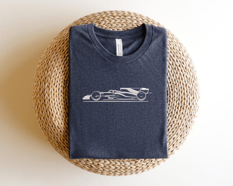 May include: A dark grey t-shirt with a white outline of a racing car on the front. The t-shirt is folded and sitting on a woven straw mat.