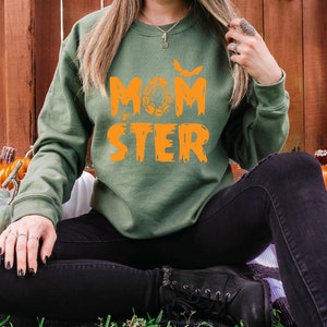K&ouml;nnte beinhalten: Ein gr&uuml;nes Sweatshirt mit dem Text "MOMSTER" in orangefarbenen Buchstaben. Das Sweatshirt hat ein Fledermaus- und Spinnennetz-Design. Das Model tr&auml;gt eine schwarze Jeans und sitzt auf einem Stapel K&uuml;rbisse.