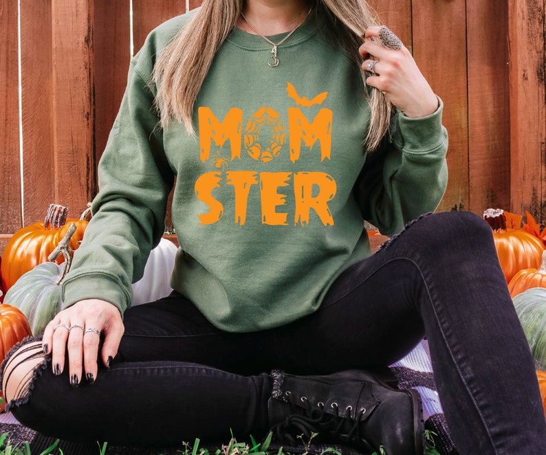 K&ouml;nnte beinhalten: Ein gr&uuml;nes Sweatshirt mit dem Text "MOMSTER" in orangefarbenen Buchstaben. Das Sweatshirt hat ein Fledermaus- und Spinnennetz-Design. Das Model tr&auml;gt eine schwarze Jeans und sitzt auf einem Stapel K&uuml;rbisse.