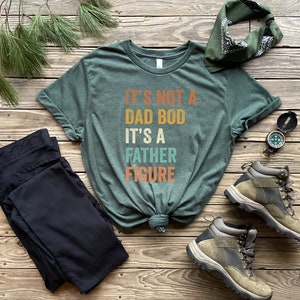 Pu&ograve; includere: Una maglietta verde con il testo "It's not a dad bod it's a father figure" in un font retr&ograve;. La maglietta &egrave; annodata ed &egrave; circondata da un paio di pantaloni neri, una bandana, una bussola e scarponi da trekking.