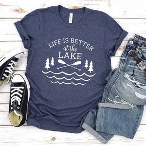 May include: A navy blue t-shirt with white text that reads "Life is better at the lake" with a graphic of two paddles crossed over each other and two pine trees on either side of the paddles. The graphic is above a wavy line representing water.