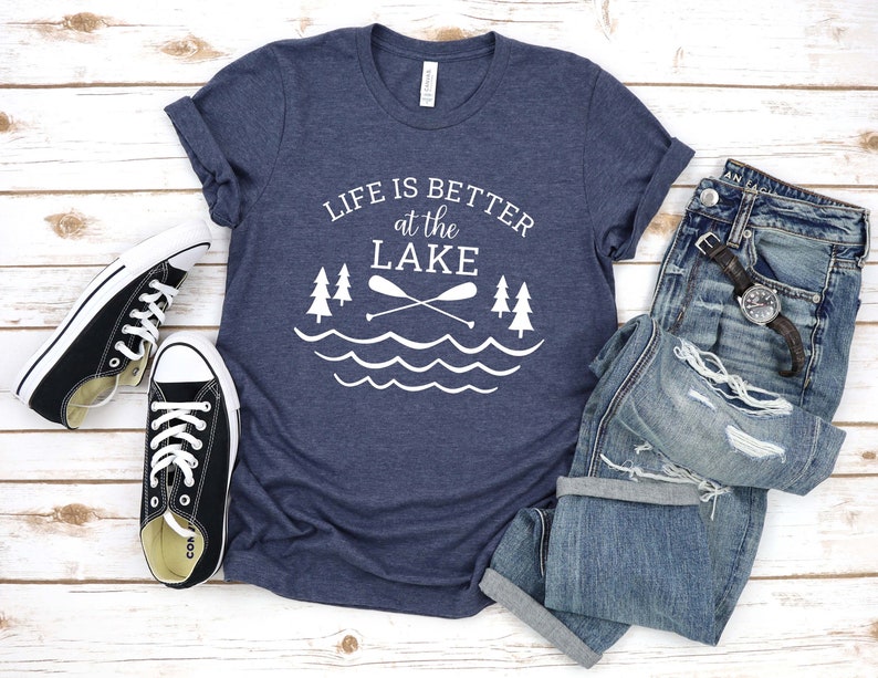 May include: A navy blue t-shirt with white text that reads "Life is better at the lake" with a graphic of two paddles crossed over each other and two pine trees on either side of the paddles. The graphic is above a wavy line representing water.