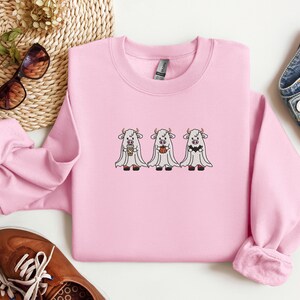 K&ouml;nnte beinhalten: Rosafarbener Pullover mit einem Motiv von drei wei&szlig;en K&uuml;hen, die als Geister verkleidet sind. Zwei K&uuml;he halten eine Tasse Kaffee und einen K&uuml;rbis.