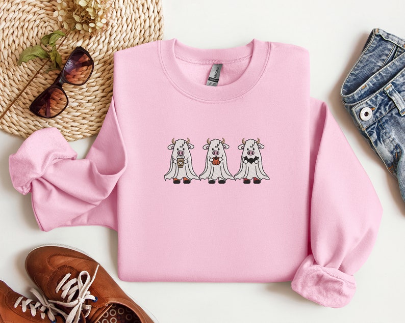 K&ouml;nnte beinhalten: Rosafarbener Pullover mit einem Motiv von drei wei&szlig;en K&uuml;hen, die als Geister verkleidet sind. Zwei K&uuml;he halten eine Tasse Kaffee und einen K&uuml;rbis.