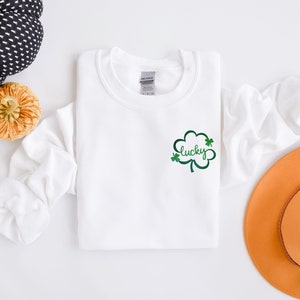 以下が含まれることがあります： 緑のクローバーのデザインと「lucky」という文字が前面に刺繍された白いスウェットシャツ
