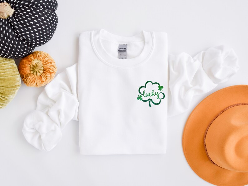 以下が含まれることがあります： 緑のクローバーのデザインと「lucky」という文字が前面に刺繍された白いスウェットシャツ