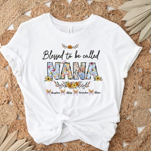 Op de afbeelding: Wit T-shirt met de tekst "Blessed to be called NANA" in zwart met bloemmotieven. De namen "Sophia, Alex, Braden, Olive" staan in een kleiner lettertype onder de tekst.