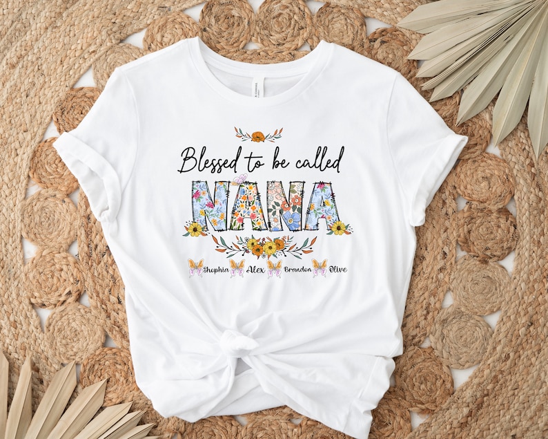 Op de afbeelding: Wit T-shirt met de tekst "Blessed to be called NANA" in zwart met bloemmotieven. De namen "Sophia, Alex, Braden, Olive" staan in een kleiner lettertype onder de tekst.