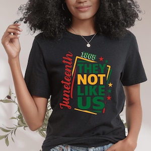 May include: Black t-shirt with the text "Juneteenth" in red script on the left side. The shirt features the text "1865 THEY NOT LIKE US" in green, yellow, and red, with star accents, celebrating Juneteenth.