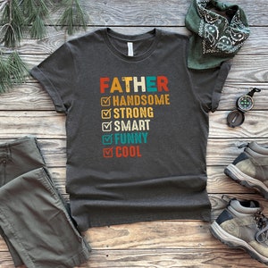 K&ouml;nnte beinhalten: Ein dunkelgraues T-Shirt mit dem Text "FATHER" in Wei&szlig; und einer Checkliste mit Eigenschaften wie "handsome", "strong", "smart", "funny" und "cool".