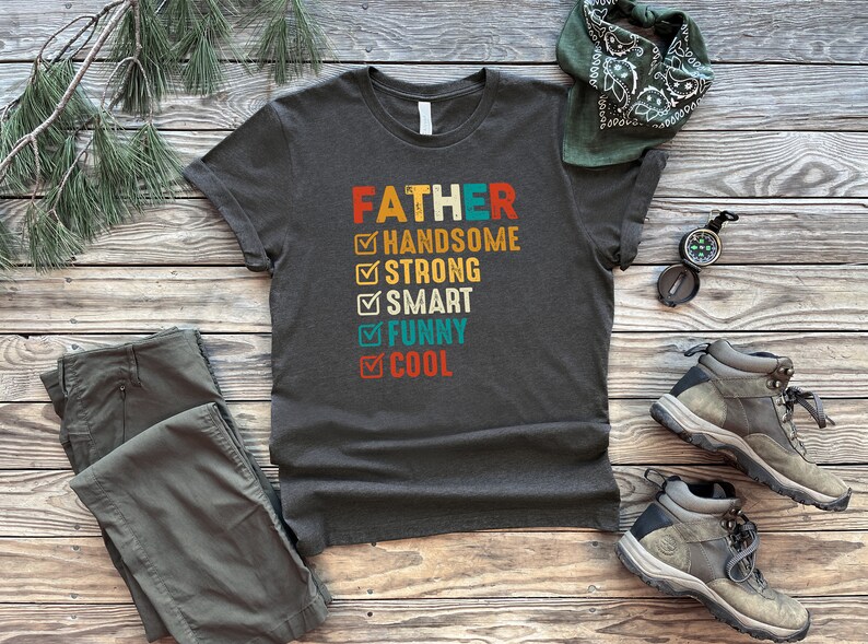 K&ouml;nnte beinhalten: Ein dunkelgraues T-Shirt mit dem Text "FATHER" in Wei&szlig; und einer Checkliste mit Eigenschaften wie "handsome", "strong", "smart", "funny" und "cool".