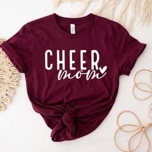 May include: Burgundy t-shirt with white text that reads "CHEER mom" with a small heart after the word "mom".