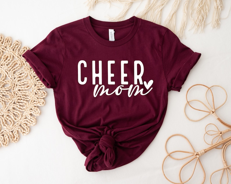 May include: Burgundy t-shirt with white text that reads "CHEER mom" with a small heart after the word "mom".